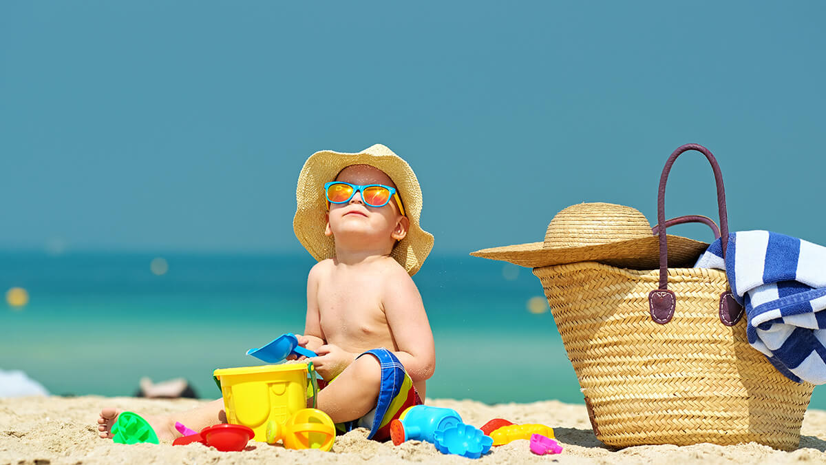A young boy sitting in the sand wearing a sun hat and sun glasses