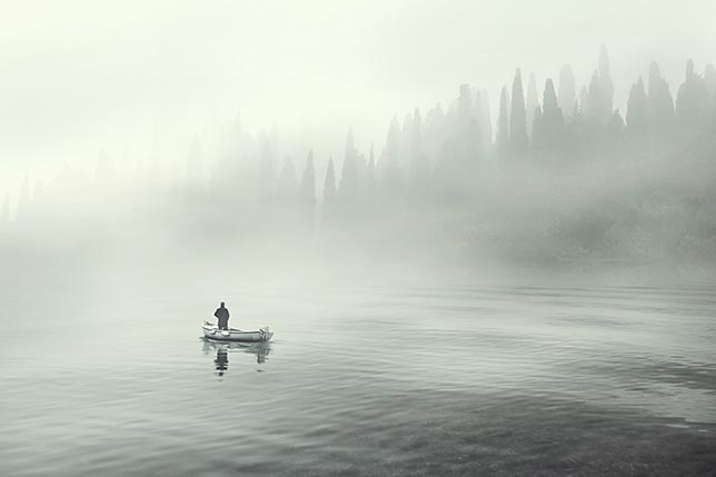 A fisherman on a small boat in the middle of a cool crisp lake as the fog slowly breaks