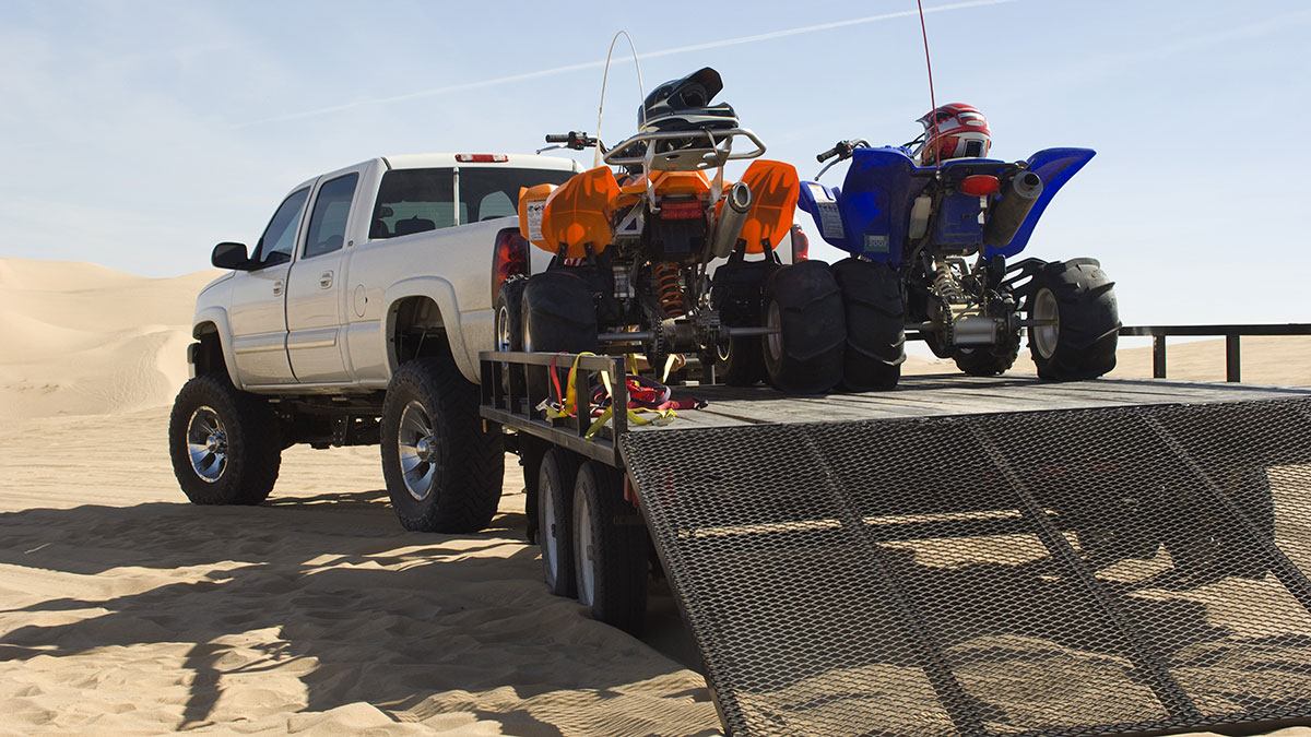 Two ATVs loaded on a trailer attached to a large white pickup truck parked amoung sand dunes. (opens in new window)