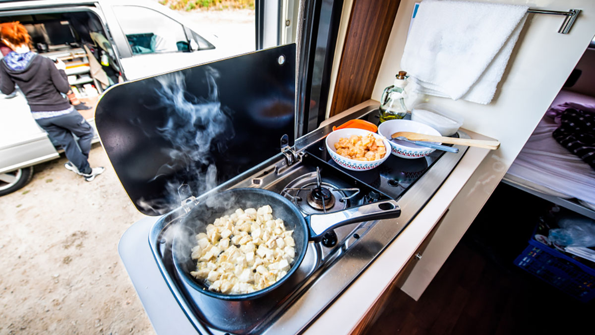 View from inside an RV of scrambled eggs cooking on stove top with window behind showing another traveler outside at their car.