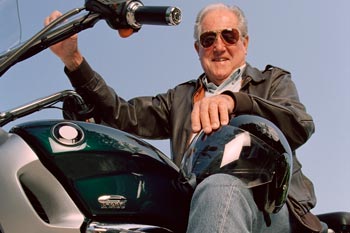 An elderly gentleman sitting on parked motorcycle with helmet on knee