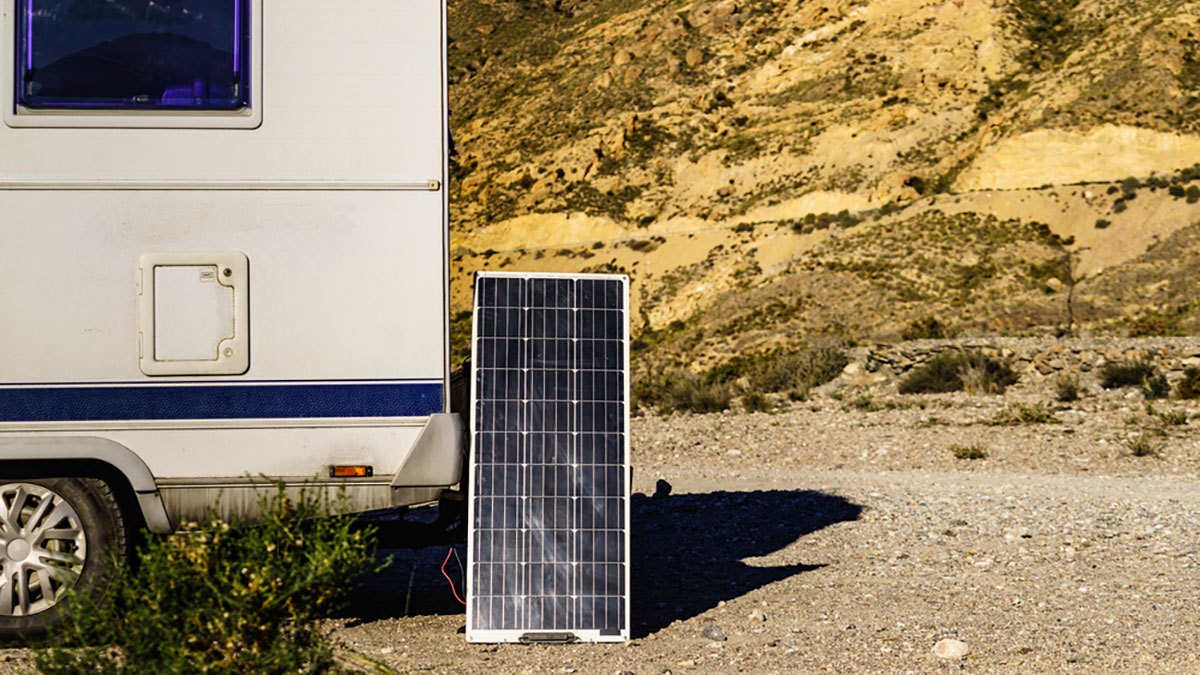 RV parked infront of an arid hillside with only the side back half of the RV in view. A solar panel rests on its bumper.